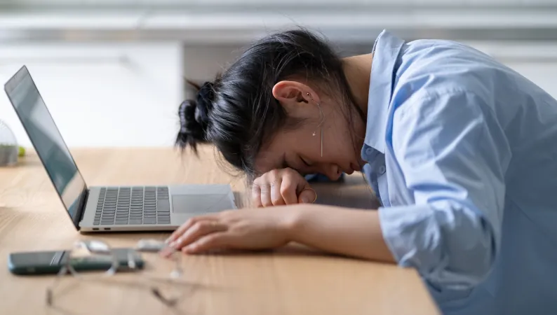 Kobieta opierająca głowę na biurku obok otwartego laptopa, wyczerpana pracą online – przykład objawu zjawiska cyfrowe zmęczenie.