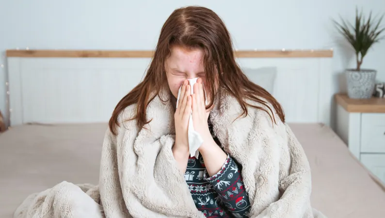 Kobieta chora na przeziębienie siedząca w łóżku pod kocem i wycierająca nos chusteczką – Reishi w sezonie przeziębień.