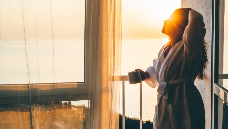 Poranny rytuał z adaptogenami – kobieta w szlafroku z kubkiem kawy lub herbaty, ciesząca się wschodem słońca i spokojnym początkiem dnia.